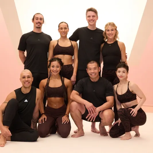 Eight people in athletic wear posing in a fitness studio with a light pink geometric background, showing off their Les Mills BodyBalance energy.