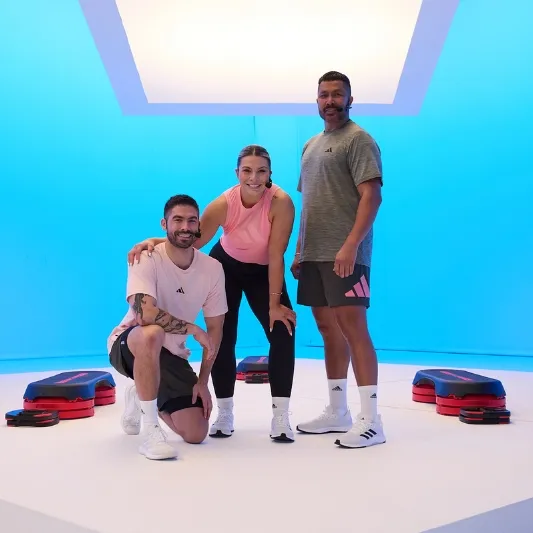 Three individuals in athletic wear posing in a brightly lit studio with a blue background, surrounded by step platforms and risers, commonly used in Les Mills BodyStep classes.