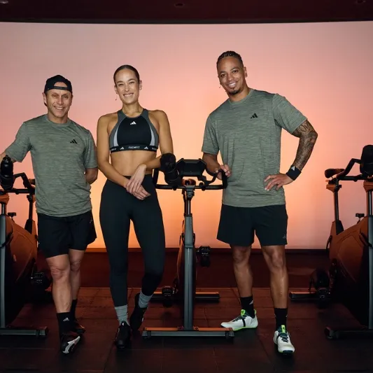 Three individuals standing next to stationary bikes in a fitness studio, wearing athletic attire, associated with Les Mills Sprint.