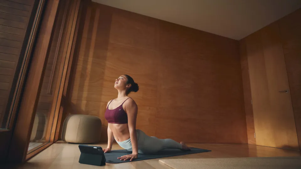 One person in a maroon sports bra and light blue leggings does yoga on a blue mat in a warm brown wooden room with large windows.