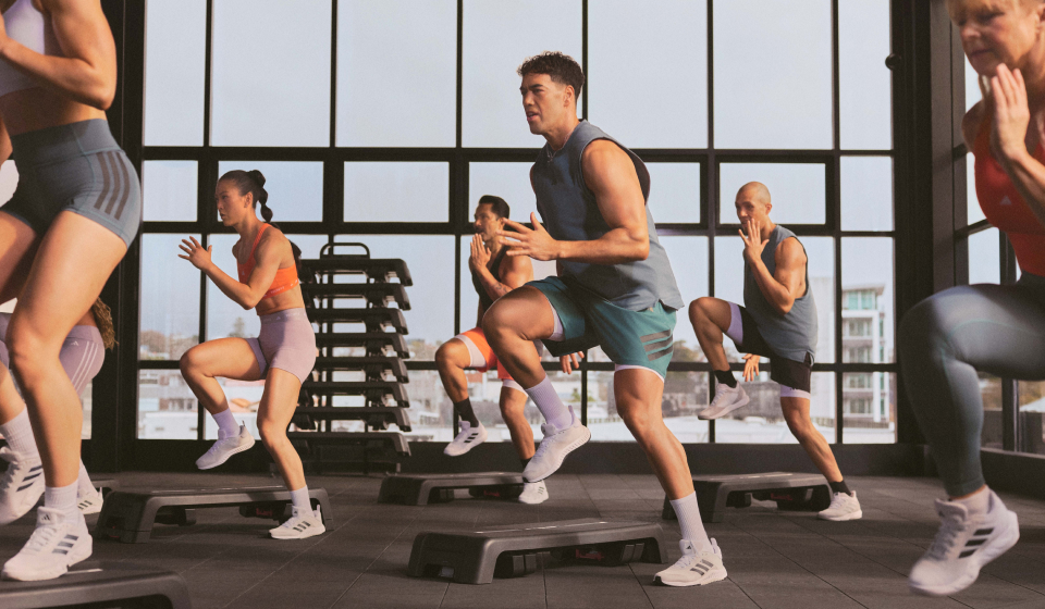 A group of people in athletic wear perform step exercises on platforms in a gym with large windows, mid-motion with one leg raised and arms balancing.