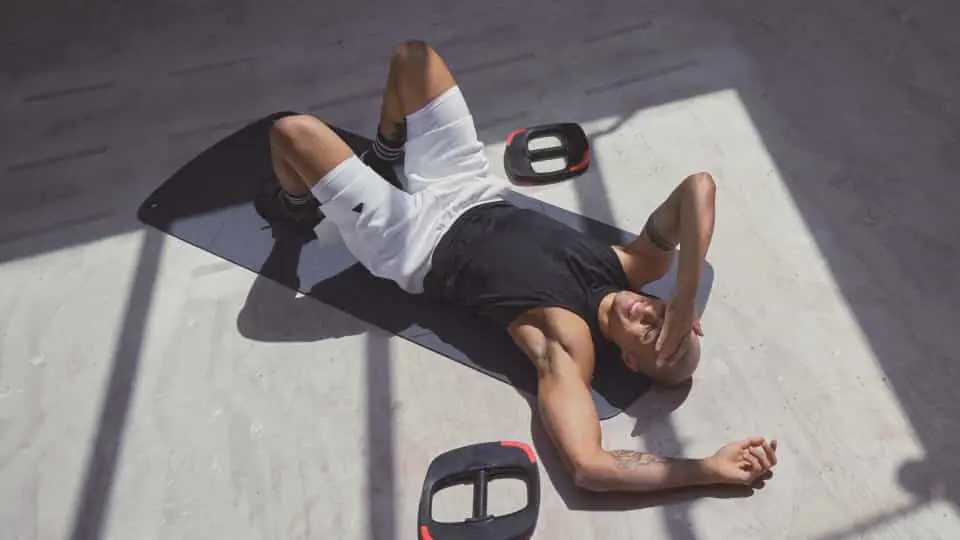 One person lying on a black exercise mat in a bright room wearing a black sleeveless top and white shorts. Two black and red weight plates are placed near the mat on the light-coloured floor. 