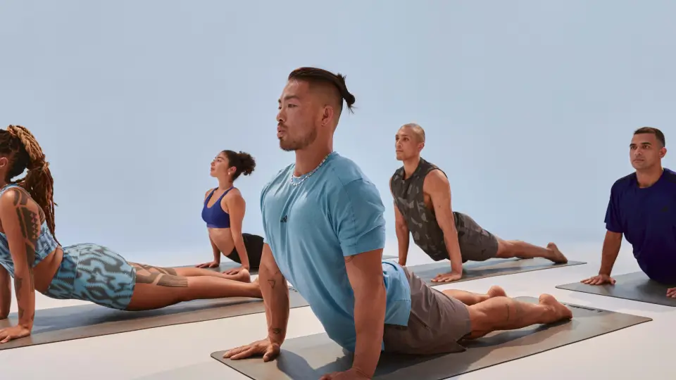 Three people exercising in a bright gym with brick walls and large windows. Foreground person wears a grey top, mint green shorts, and black shoes, stepping on a red platform while holding a barbell.