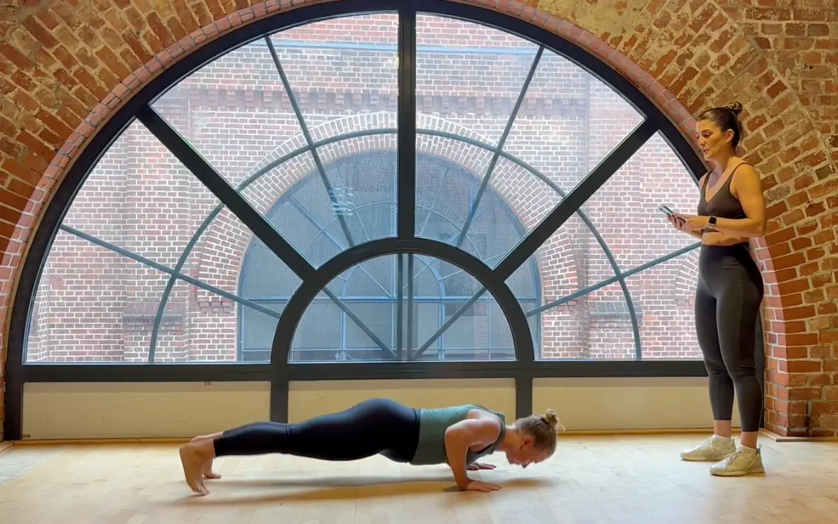 Two people in workout gear in front of a large arched window with brick framing. One is doing a push-up on a wooden floor wearing a green top and black leggings, the other stands holding a device.
