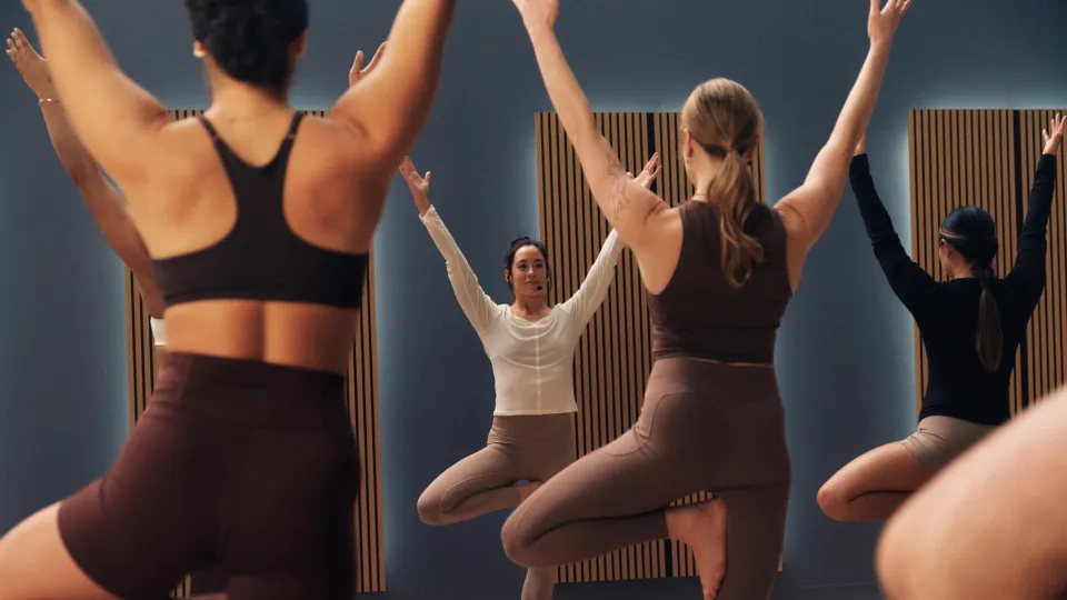 Five people in a yoga studio with wooden floors and acoustic panels, standing on dark mats in tree pose with arms raised. Outfits include brown, beige, and black workout tops and leggings.