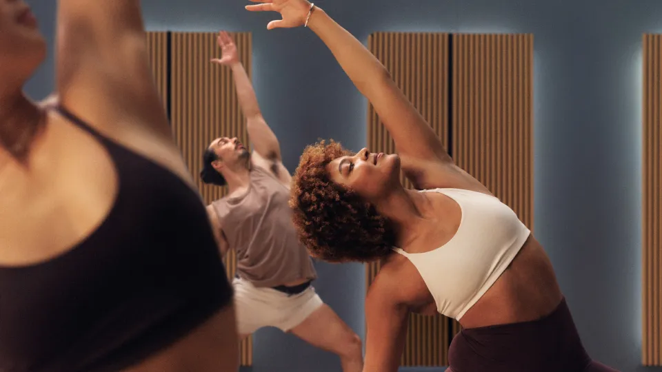 People in a yoga studio with wooden floors and acoustic panels, performing side stretch poses. Outfits include white and brown sports tops with matching leggings in a softly lit room.
