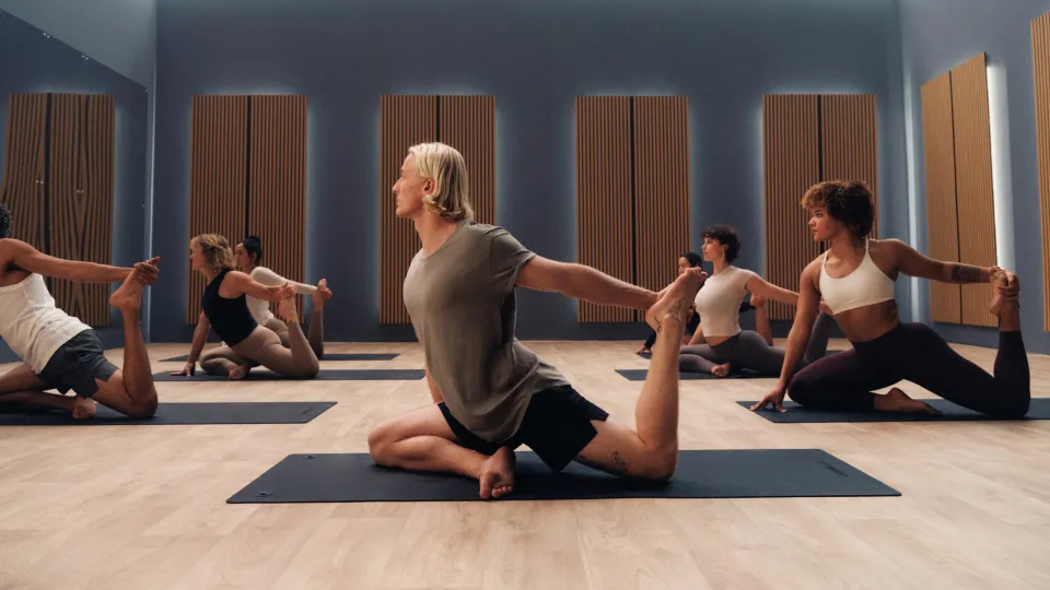 Six people in a yoga studio with wooden floors and acoustic panels, seated on dark mats in a twist pose holding one foot behind them. Outfits include grey, white, and brown workout tops and leggings.