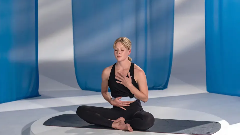 Person seated cross-legged on a black yoga mat in a bright studio with blue curtains, wearing a black workout top and leggings, with one hand on the chest and the other on the abdomen.