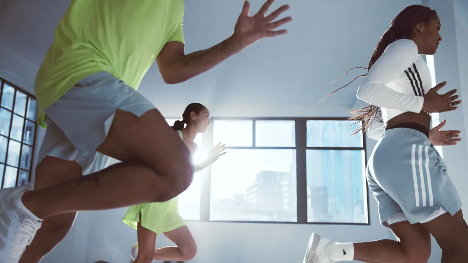 Three people running indoors near large windows. One in neon green top and shorts, another in white long-sleeve with grey shorts, third in bright green.