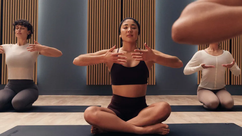 Four people seated on yoga mats in a studio with wooden panels. One wears a dark brown sports bra and shorts, others in light tops and grey leggings, arms raised in a breathing pose.