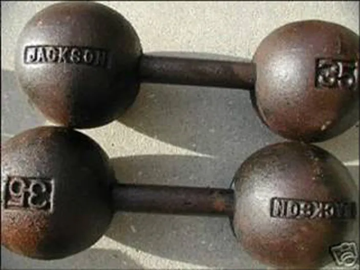 Two vintage globe-style dumbbells made of dark metal with a rusty texture, featuring engraved text “JACKSON” and weight markings “35” on each end, placed on a light concrete surface.