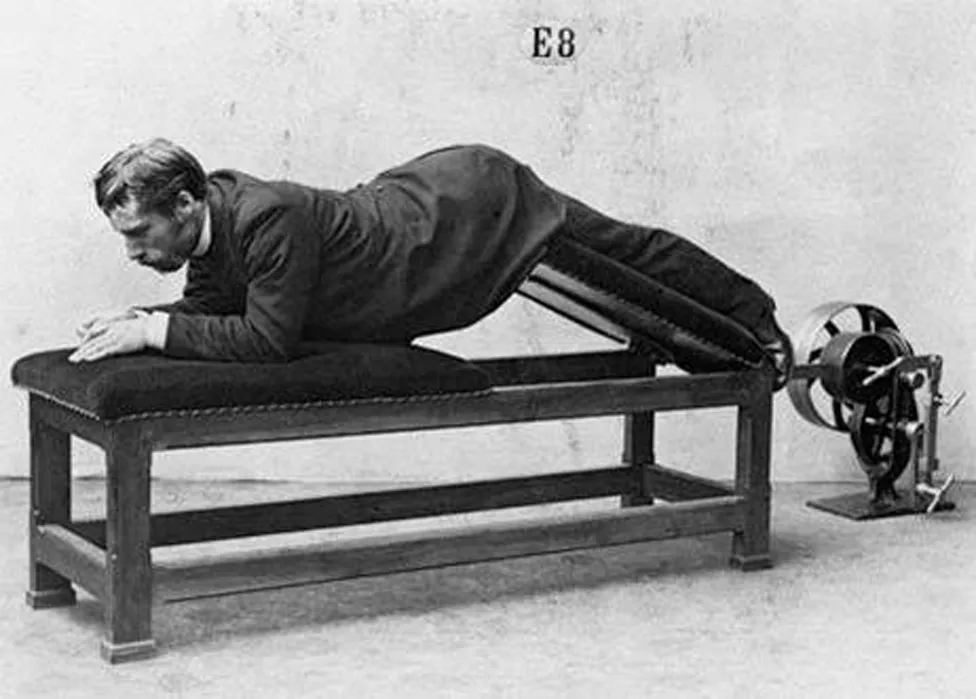 Person lying face down on a padded bench wearing a dark long coat and striped trousers, with legs secured in a mechanical device featuring metal components and a wheel on the right side.