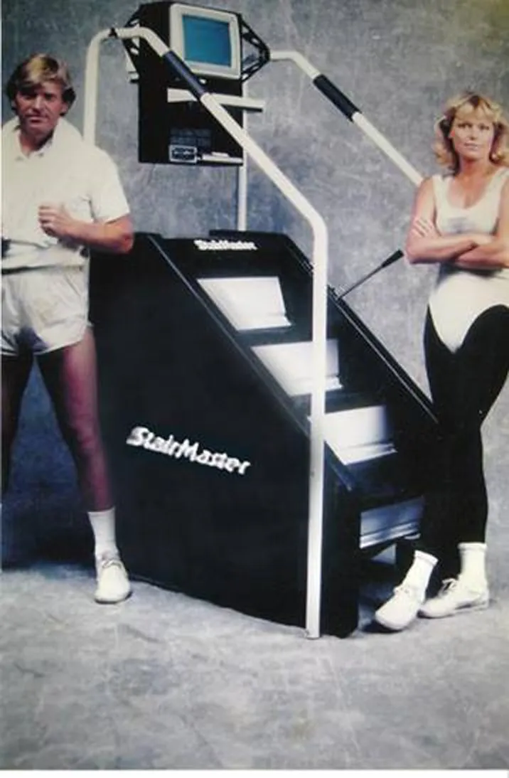 Two people standing beside a vintage black StairMaster exercise machine with white railings and a small monitor on top. They wear white and black workout outfits with white shoes against a grey backdrop.