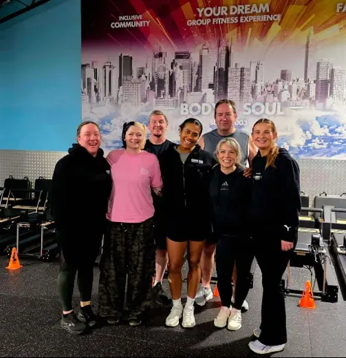 Seven people standing in a gym with black flooring and orange cones. Behind them is a large wall mural showing a city skyline with bright pink, orange, and white colours and fitness-themed text.