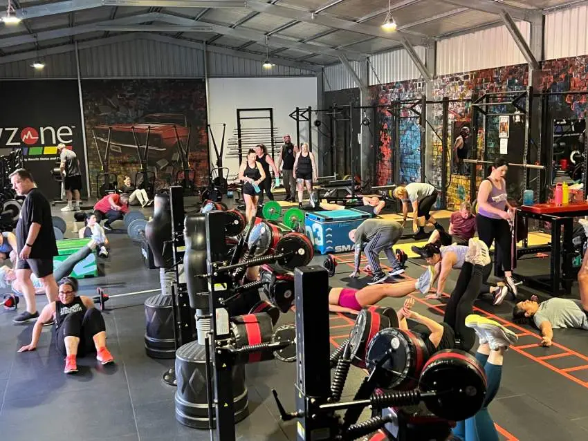 Indoor gym with about 18 people doing functional training on black flooring. Equipment includes bikes, barbells, and green boxes. Colourful graffiti wall and metal roof visible in background.