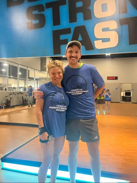 Two people in blue T-shirts and shorts standing on wooden flooring in a gym. Behind them is a mirrored wall and a large blue sign with bold black and white text reading “STRONG FAST.”