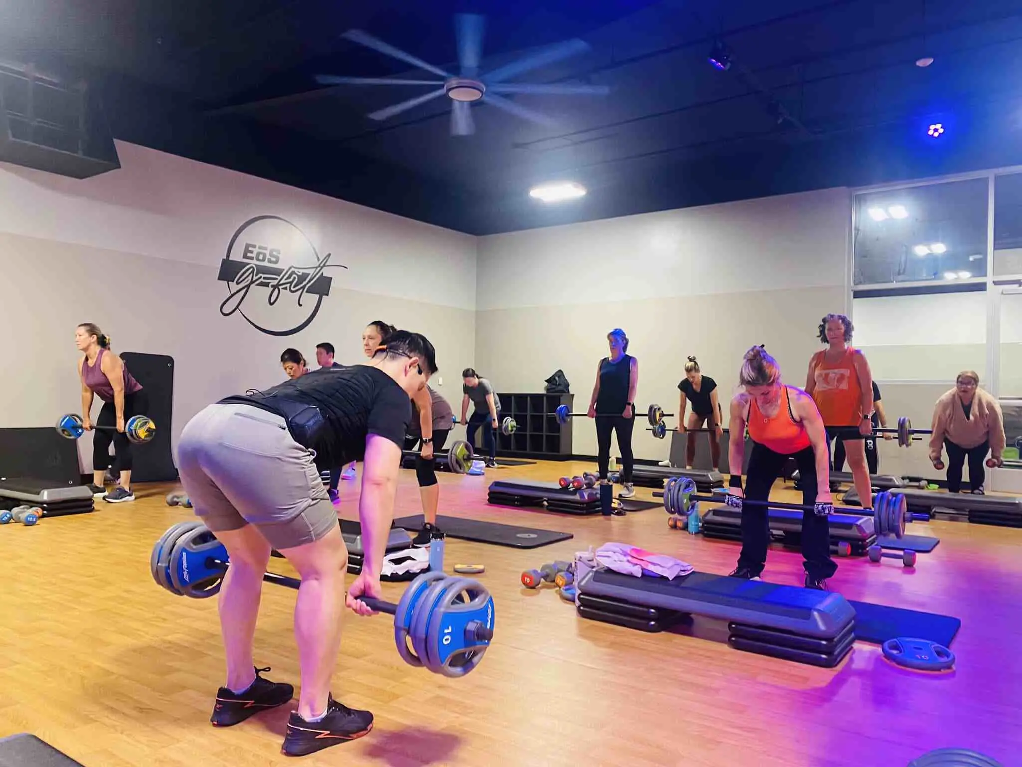 Indoor gym with 12 people lifting barbells on black steps. Wooden flooring, light grey walls, and dark ceiling with bright lights. Blue and orange weight plates scattered around the studio.