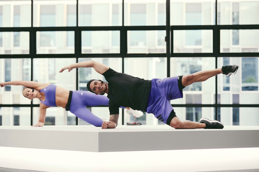 Two people performing side plank variations on a raised platform in a bright studio with large windows.