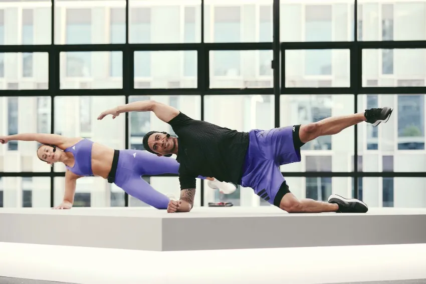 Two people performing side plank variations on a raised platform in a bright studio with large windows.