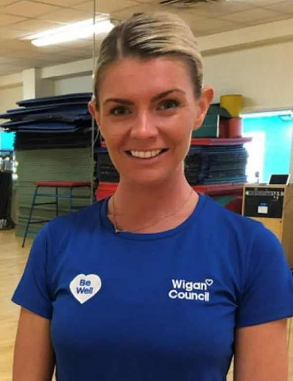 Smiling woman wearing a blue Wigan Council shirt with 'Be Well' logo standing indoors in a gym or fitness setting.
