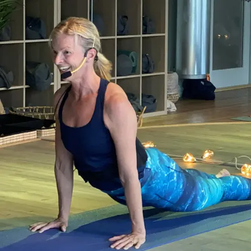  Person in athletic clothing holding a forward‑leaning yoga pose on a round red‑lit stage, with others exercising behind them in a dim studio with wooden floors and string lights.