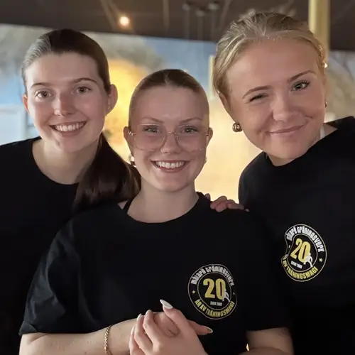 Three people standing closely together wearing matching black T‑shirts with a circular gold “20” anniversary logo in an indoor setting.