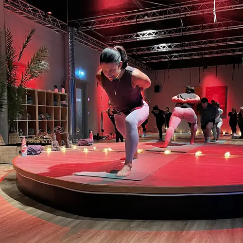 Person in athletic clothing holding a forward‑leaning yoga pose on a round red‑lit stage, with others exercising behind them in a dim studio with wooden floors and string lights.