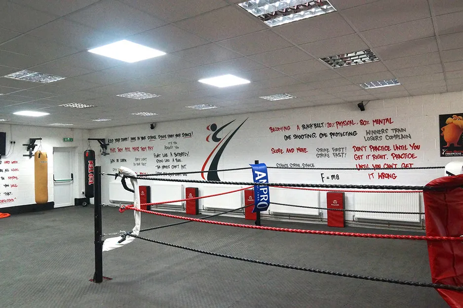 Empty boxing gym with a boxing ring in the center, punching bags hanging on the left wall, and motivational quotes written on the back wall.