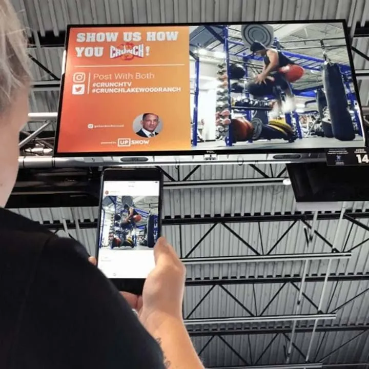 Person taking a photo with a smartphone of a gym TV screen showing a person climbing gym equipment and a social media call to action for #CrunchTV and #CrunchLakewoodRanch.