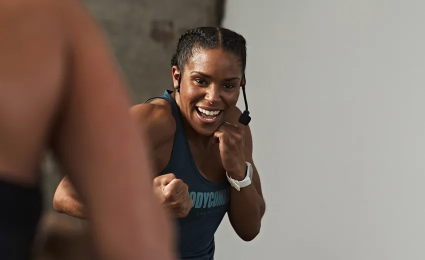 Female fitness instructor wearing a headset microphone and tank top, demonstrating a boxing stance with a focused smile.