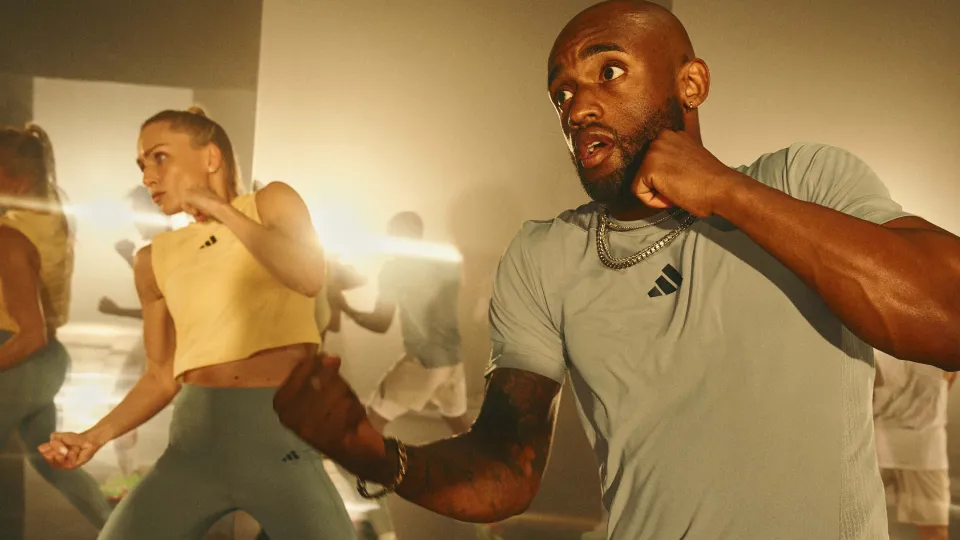 Two people in athletic clothing practicing boxing moves in a gym with a bright light behind them.
