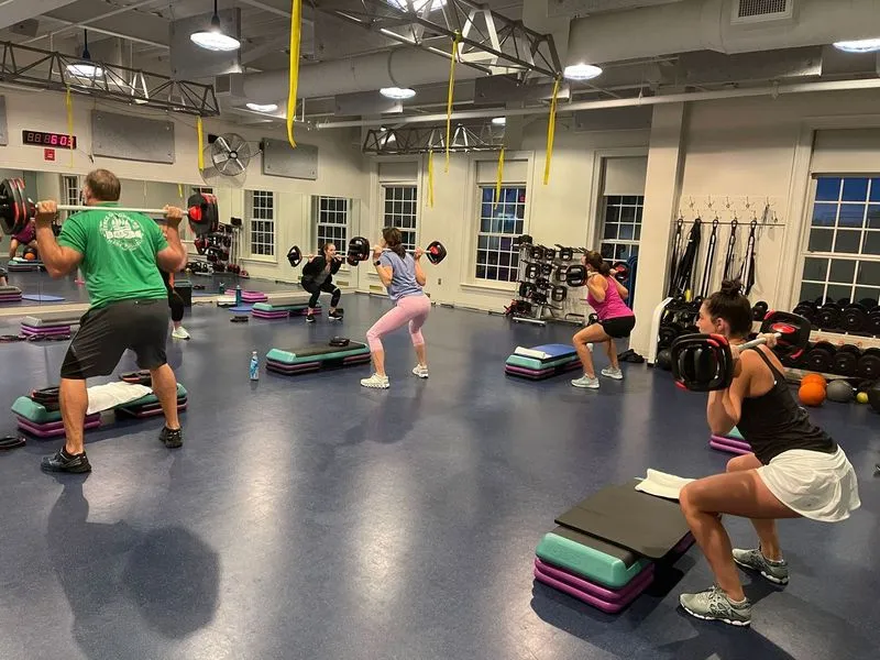 Group fitness class with five people performing squats while holding barbells in a gym with large windows.