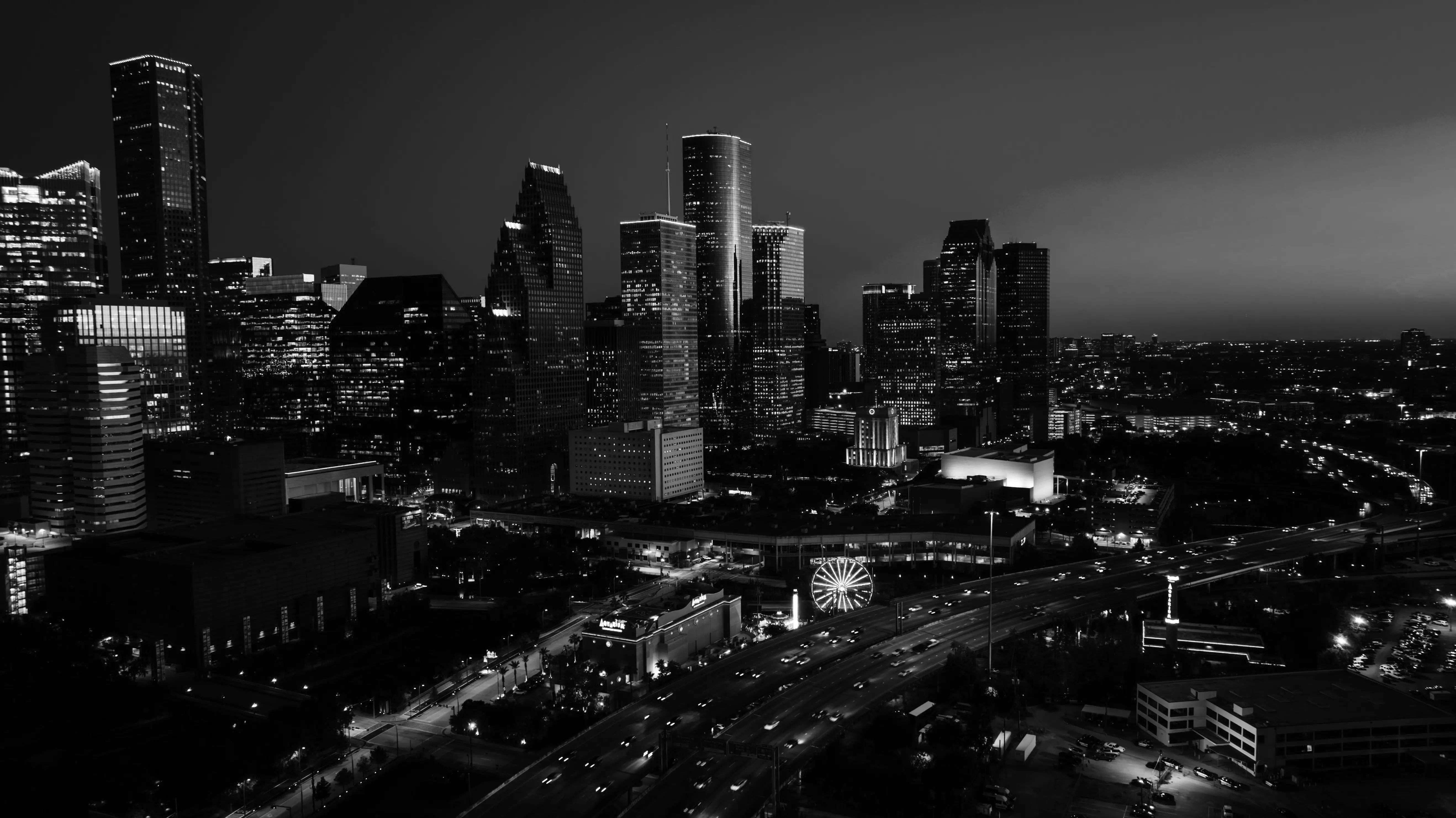 Nighttime city skyline with illuminated skyscrapers and a highway with moving vehicles in the foreground.