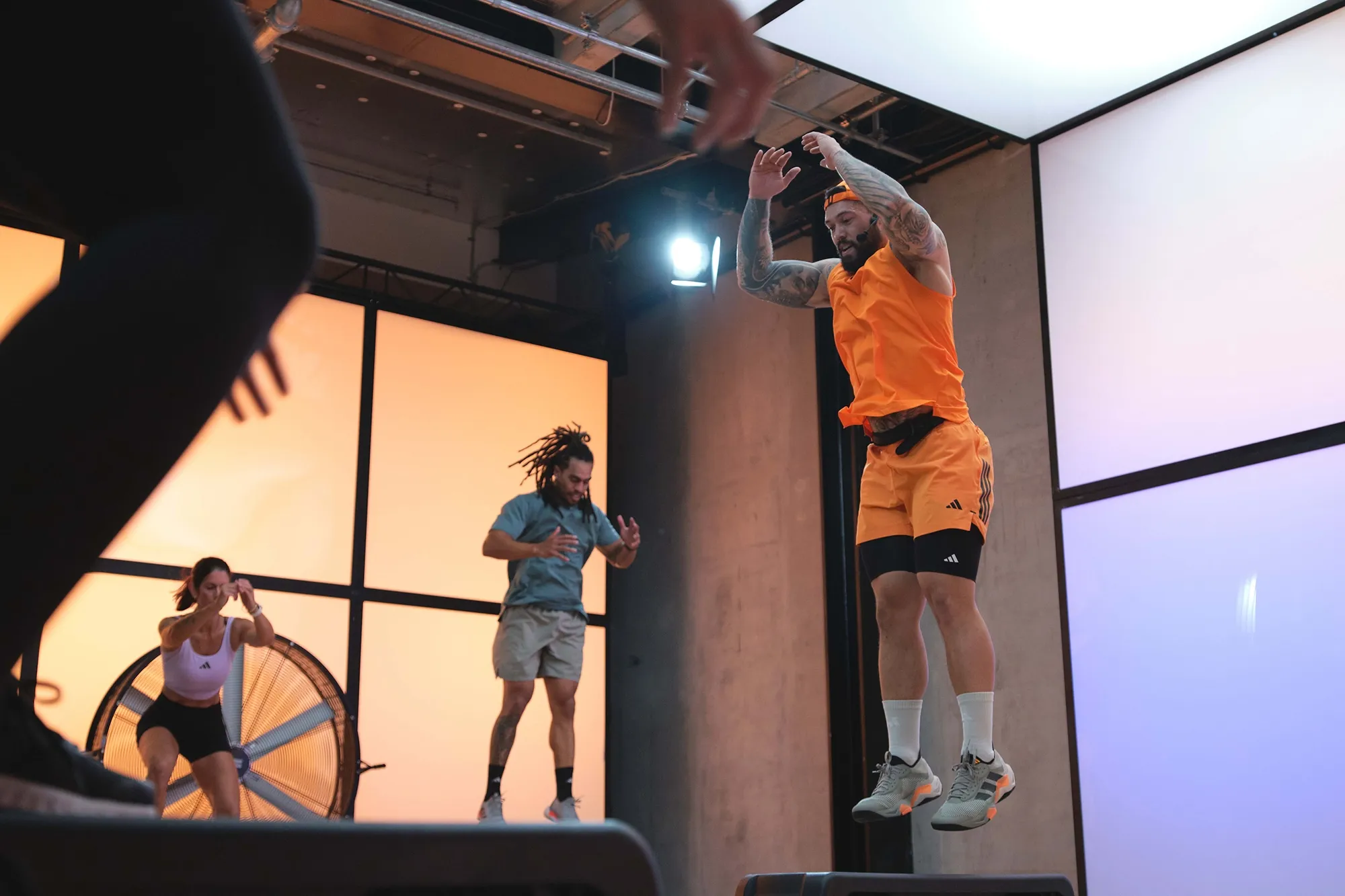 Three people performing a group fitness workout inside a studio with orange and blue backlit panels, one man jumps on a step platform.