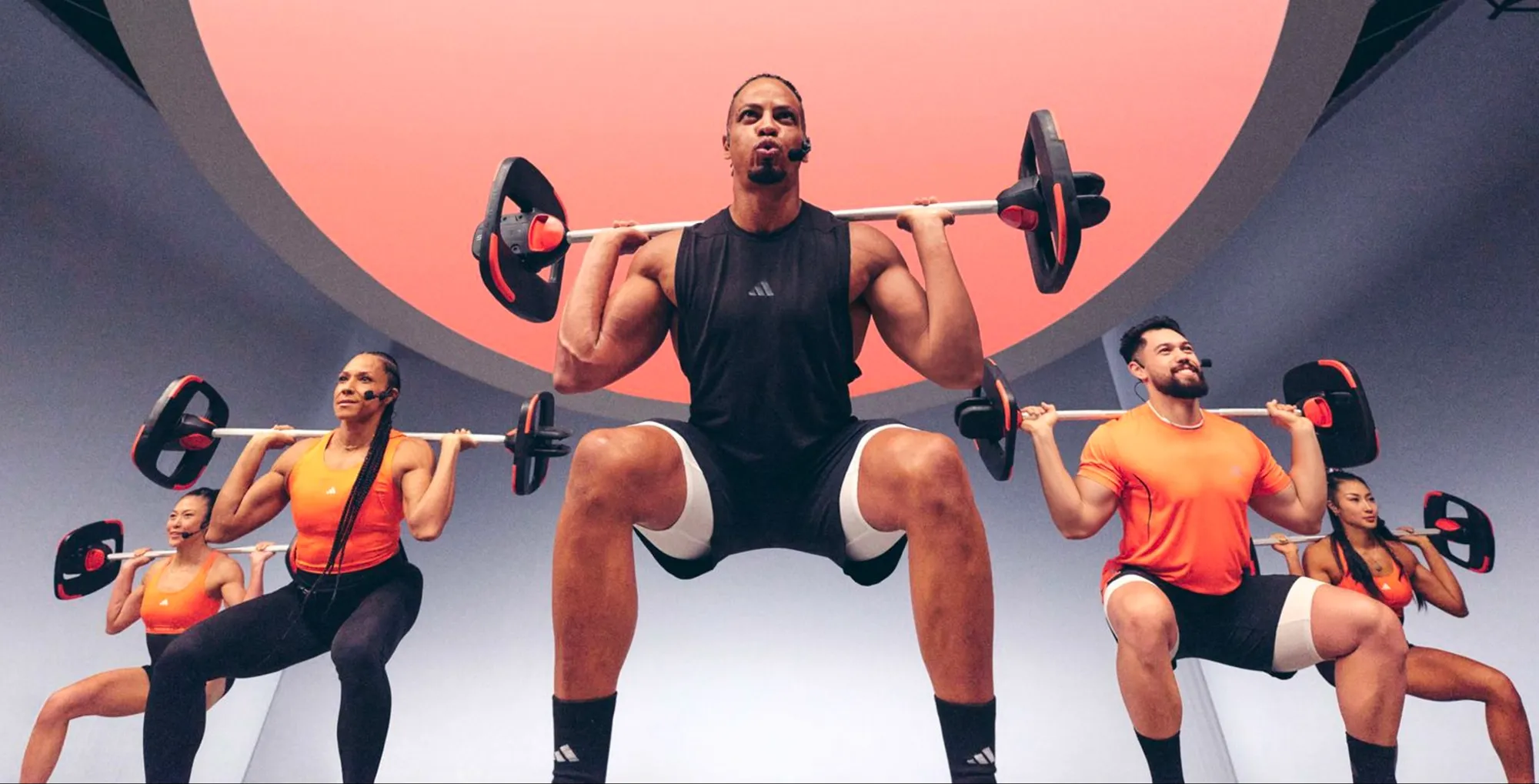 Group of five people performing barbell squats in a brightly lit, modern fitness studio with a circular pink ceiling.