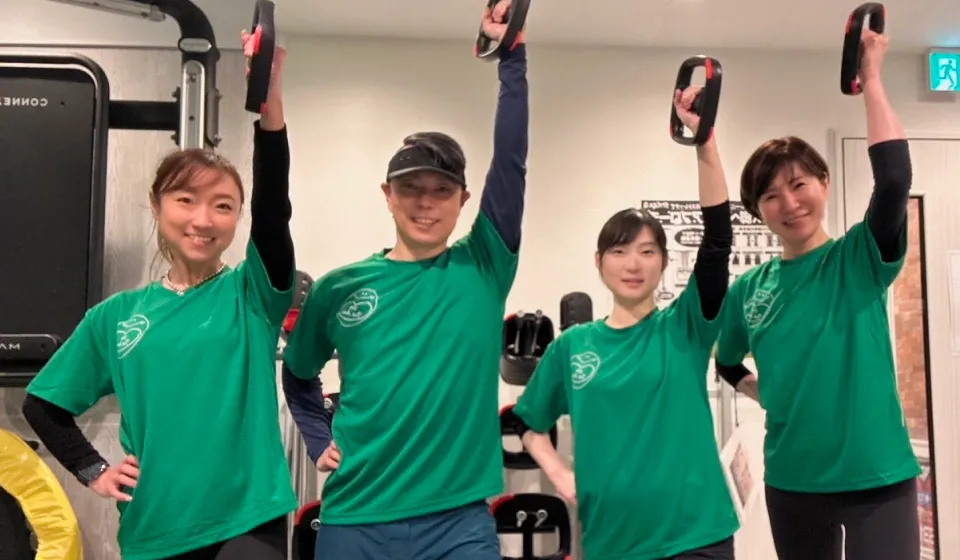 Four smiling people in green shirts holding one arm up gripping black and red fitness ring handles in a gym.