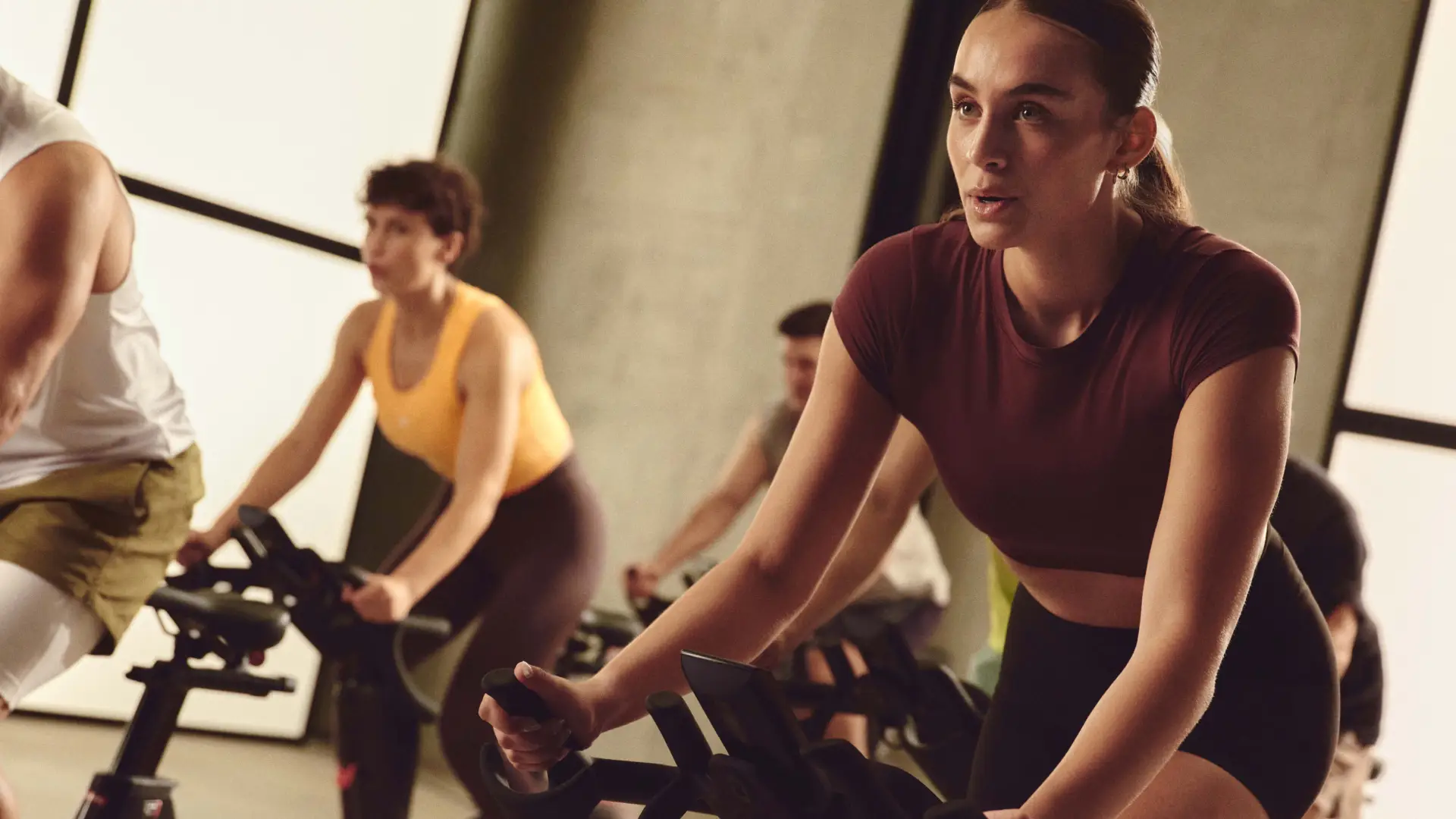 Group of people cycling indoors in a fitness class, focused on a woman in a maroon top.