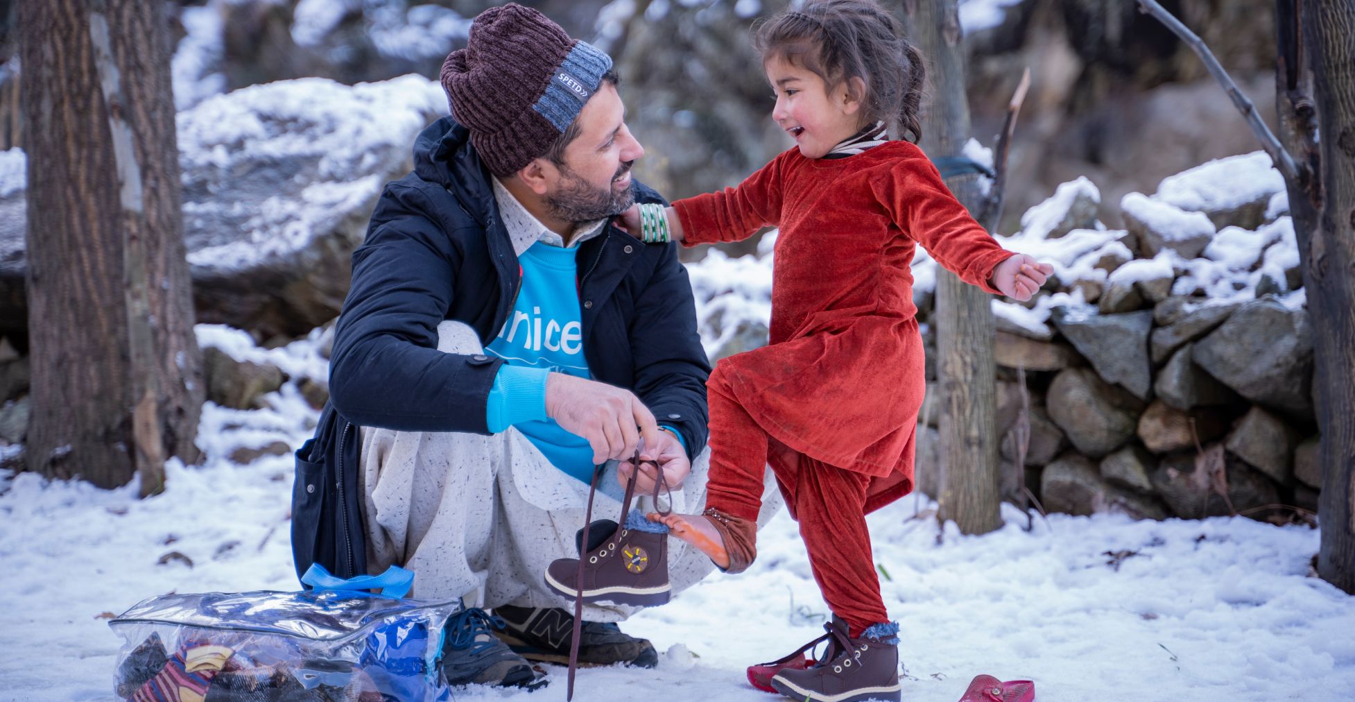 Man kneeling in snow helping a smiling girl put on her boots outdoors.