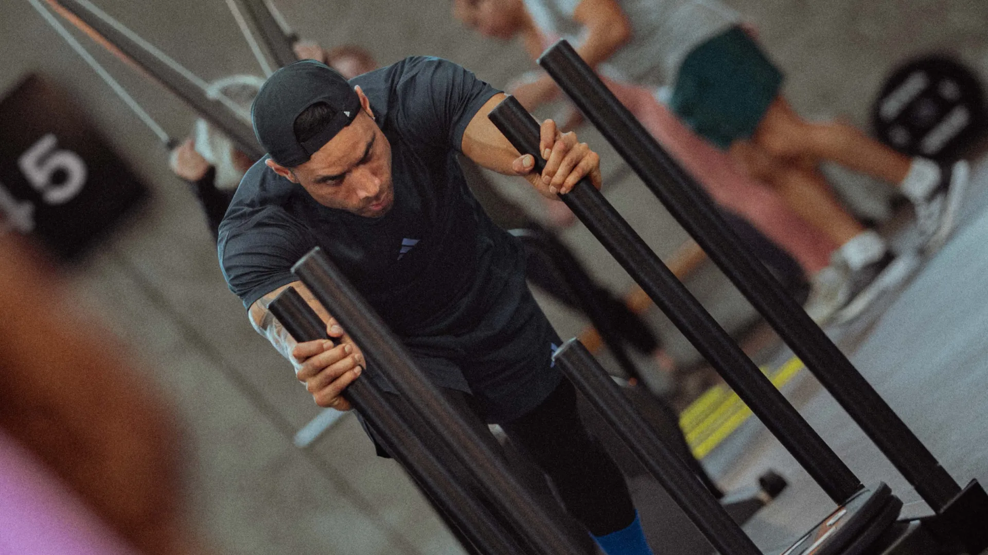 Man wearing a black cap pushing weighted sled in a gym with others exercising in the background.