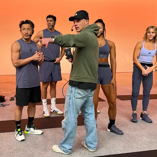 Fitness instructor in a green hoodie directs four athletes in athletic wear on a stage with an orange backdrop.