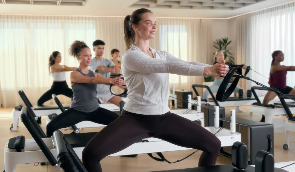 Group of people doing Pilates exercises on reformer machines in a bright, spacious studio.