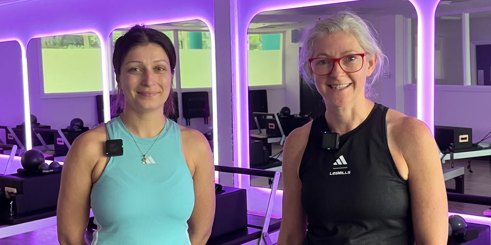 Two smiling women in workout gear stand in a fitness studio with purple neon lights and Pilates reformers.