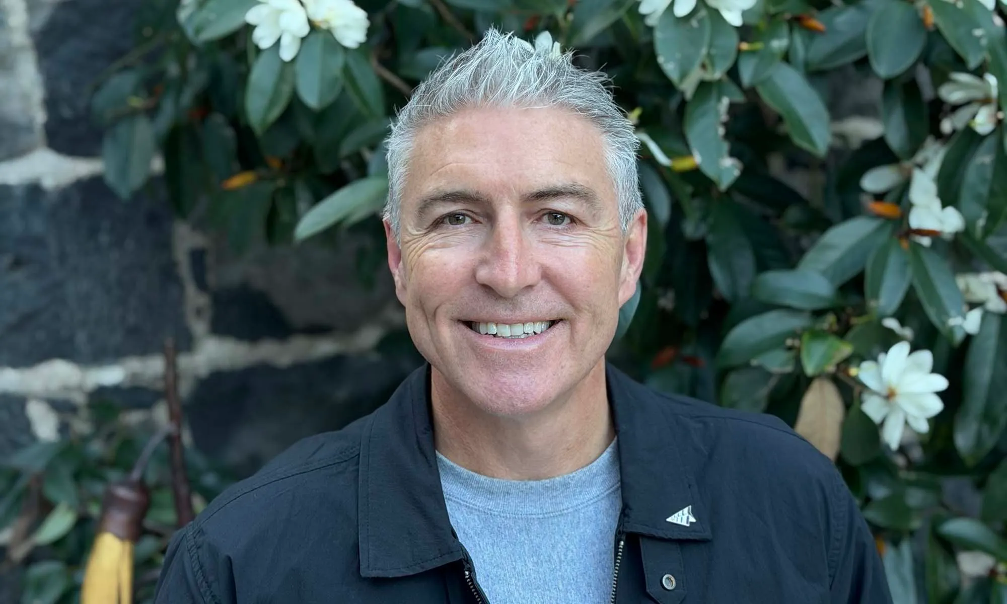 Smiling man with gray hair wearing a black jacket and gray shirt in front of leafy background.