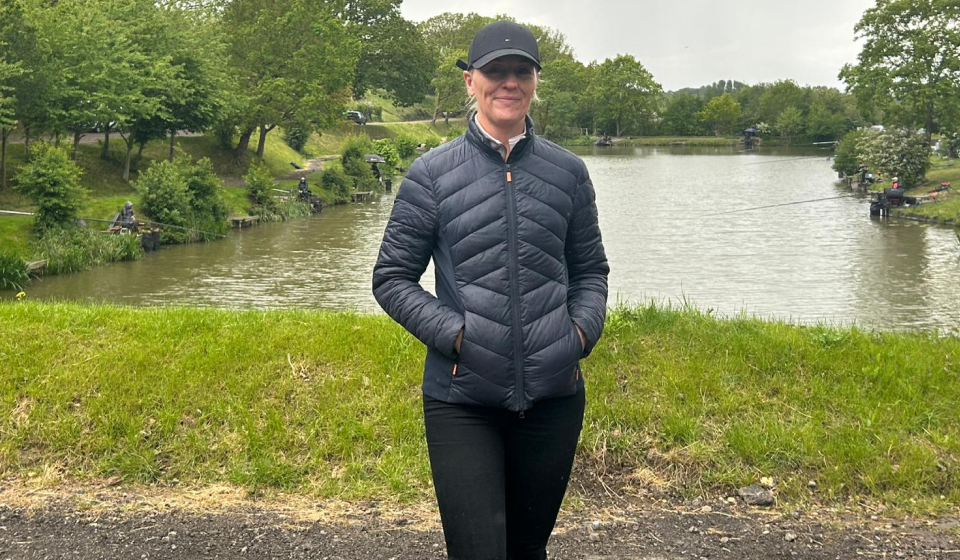 A woman in black puffer jacket, black cap stands riverside