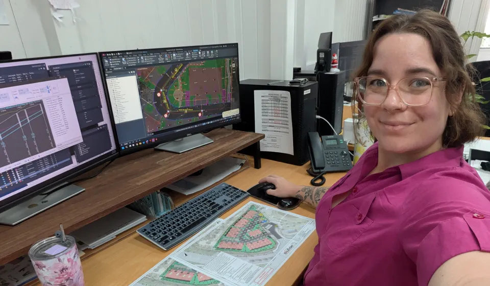 A woman with glasses in a pink shirt working at dual computer screens