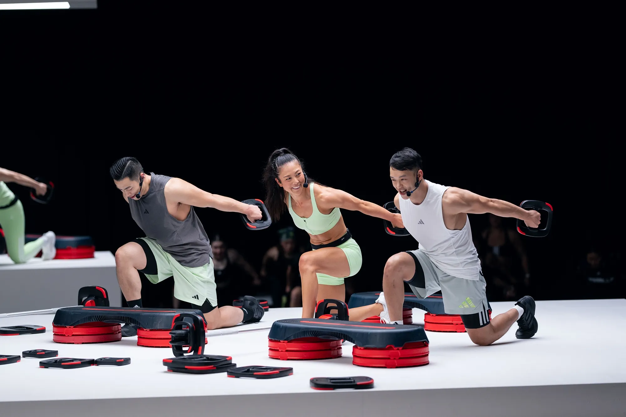Three people in athletic wear performing kneeling side raises using hand weights on a white platform with red and black step platforms around them during a fitness class.