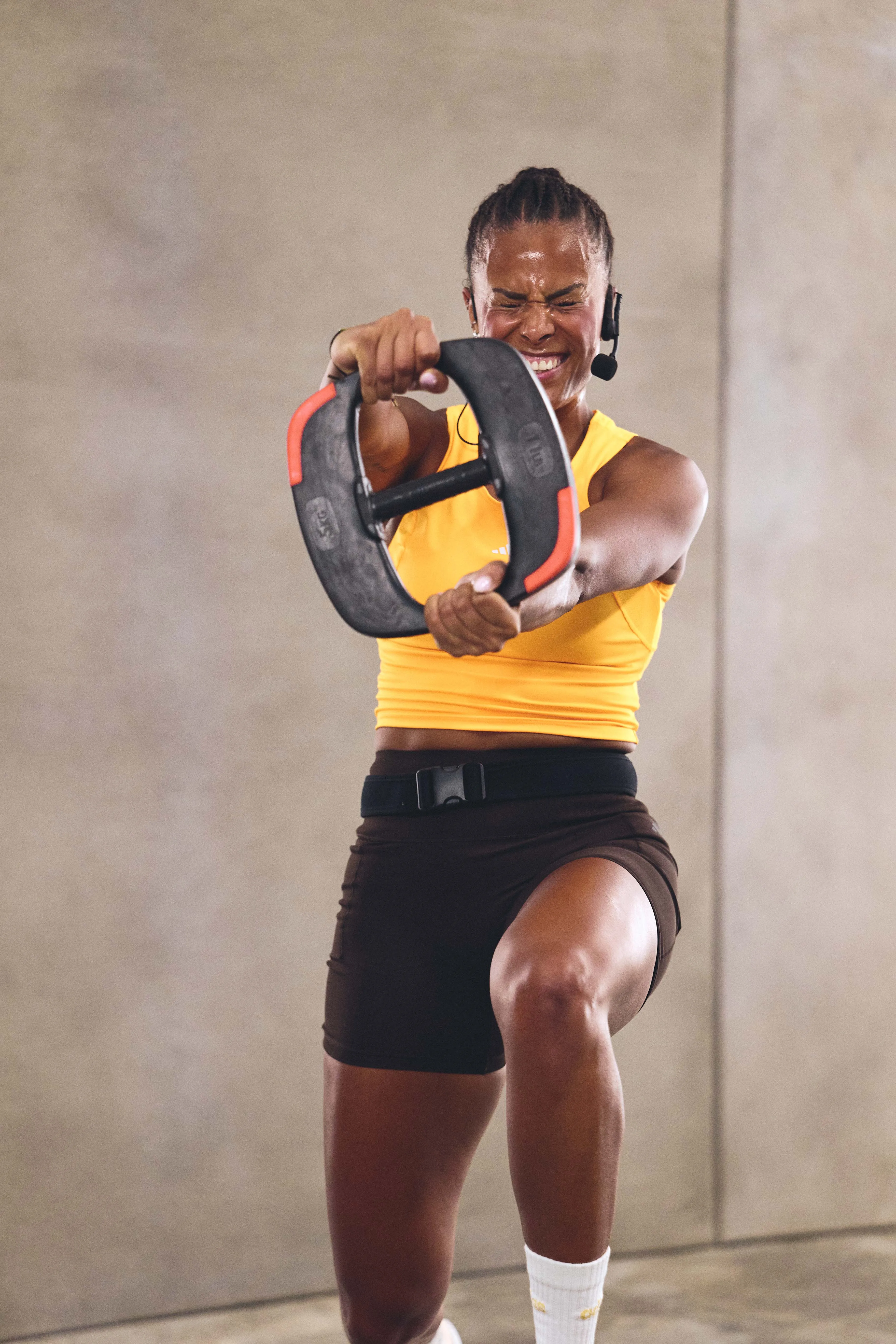 Woman in a yellow tank top and black shorts exercising indoors, holding a black and red weight plate with arms extended forward, kneeling on one leg with a focused expression and wearing a headset microphone.