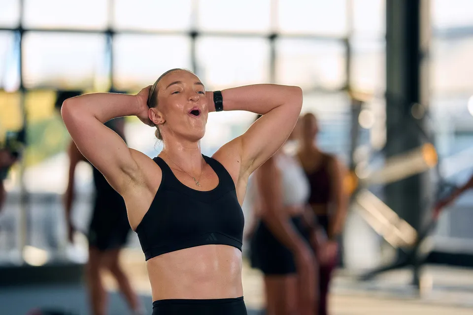 Sweaty woman in black sports bra with hands behind her head, catching breath after workout in gym with blurred people in background.