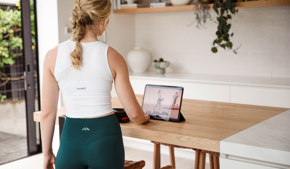 A blonde woman in white and green workout gear using an Ipad in the kitchen to watch a workout