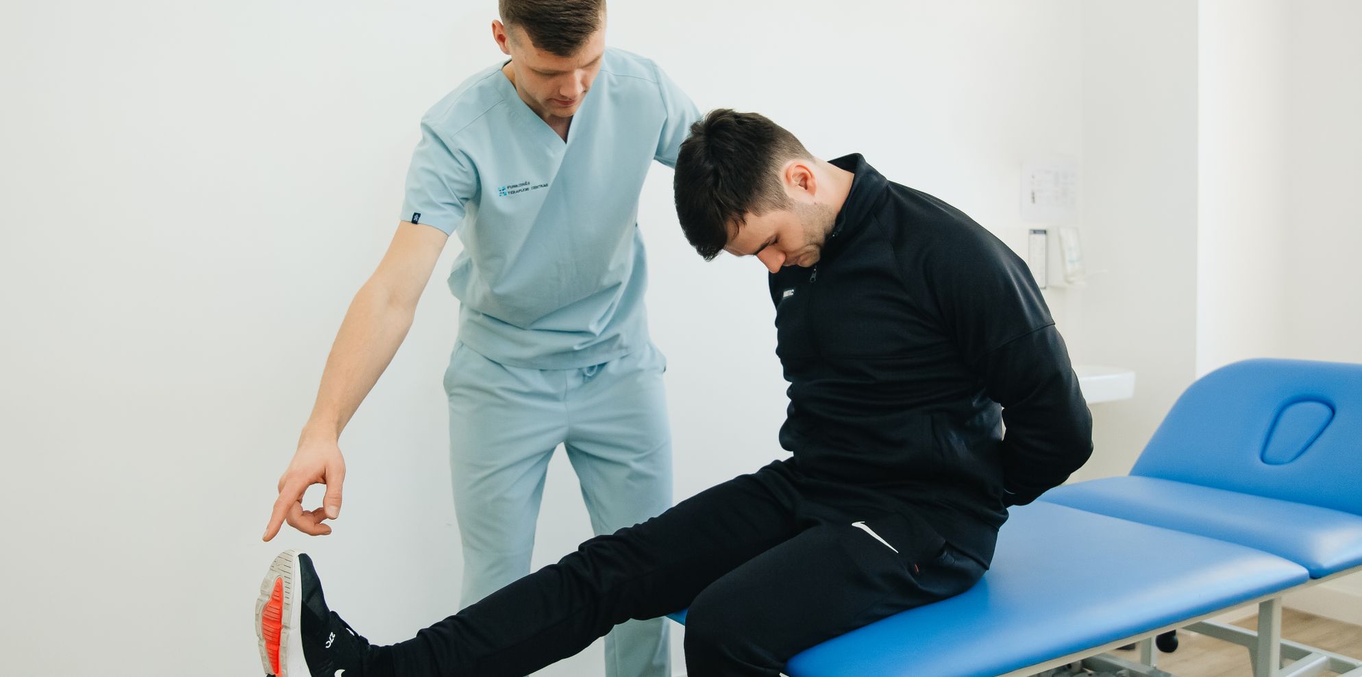 Physiotherapist in scrubs guiding a patient through a leg stretch on a clinic treatment table